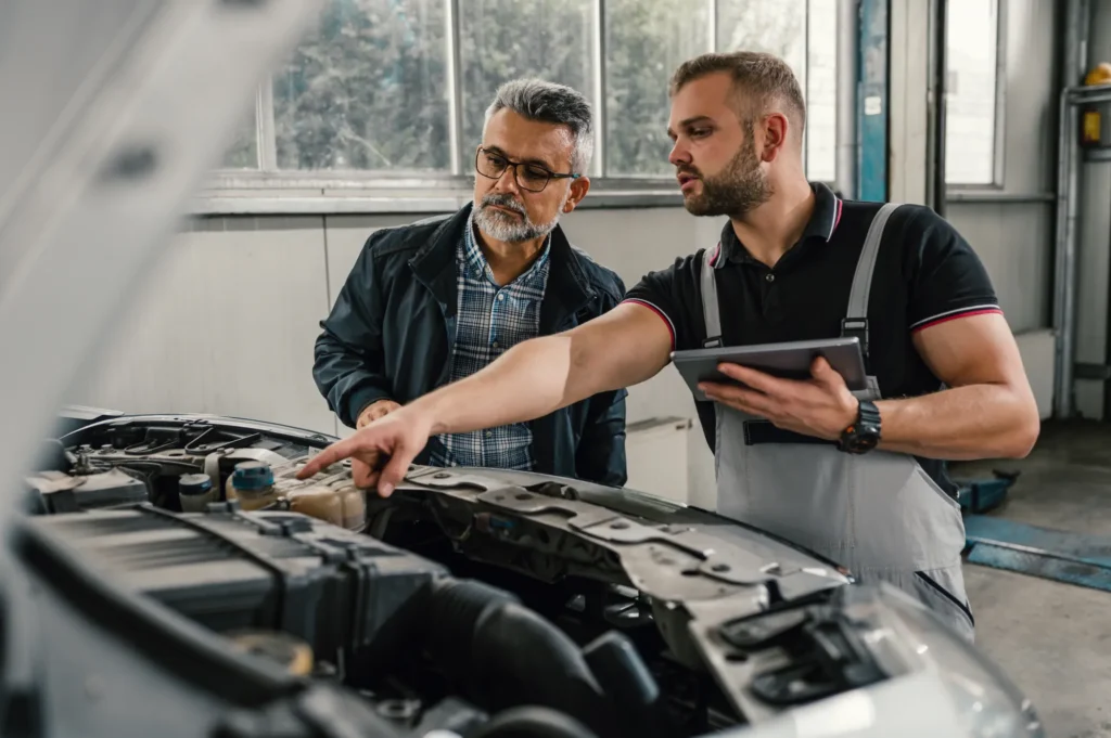 A mechanic with a tablet points at the engine under a car's open hood while explaining it to an older male customer in a repair shop.