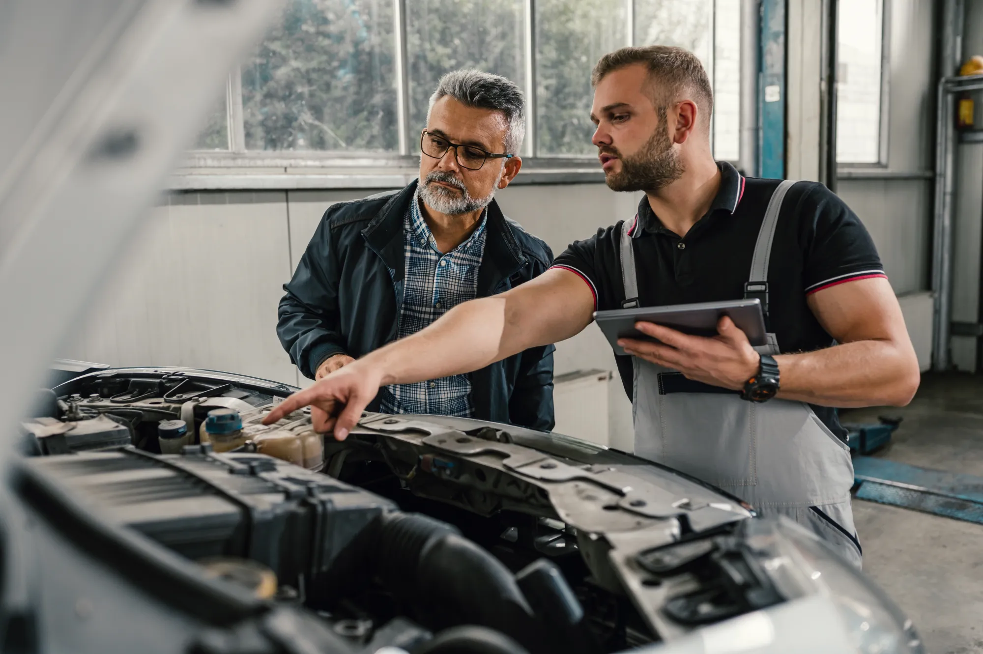 A mechanic with a tablet points at the engine under a car's open hood while explaining it to an older male customer in a repair shop.