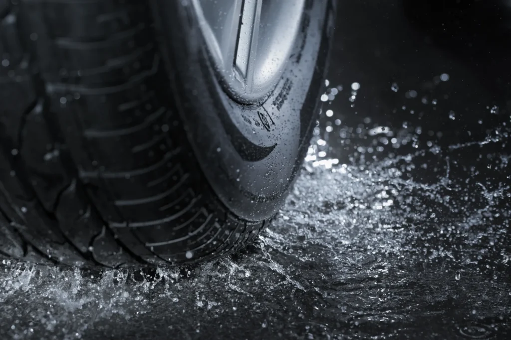 Close-up of a car tire and alloy wheel splashing through water on wet pavement.
