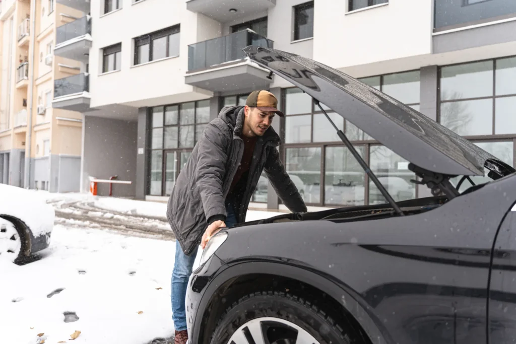 A man in a winter coat and cap leans over the open hood of a black SUV parked in the snow, looking into the engine bay.